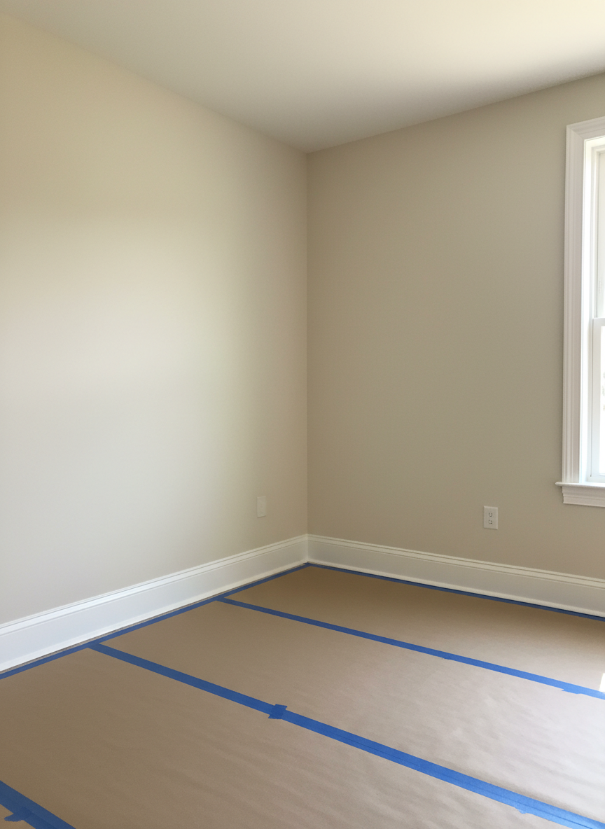 A bright, freshly painted interior room with smooth, flawless walls in a warm neutral off-white, contrasted by crisp, straight lines of pure white semi-gloss trim along baseboards, door frames, and window casings. A neatly protected hardwood floor is covered by clean rosin paper and blue painter’s tape along all edges, showing professional preparation. Natural mid-day light streams through an uncovered window, creating soft, even illumination and subtle gradients across the walls, while a faint reflection appears on the trim’s slightly glossy surface. Captured from a corner at eye level, the wide composition shows two adjoining walls and the ceiling, emphasizing uniform coverage and sharp cut lines. The photographic realism and uncluttered, modern aesthetic underscore high-quality interior painting services and attention to detail.